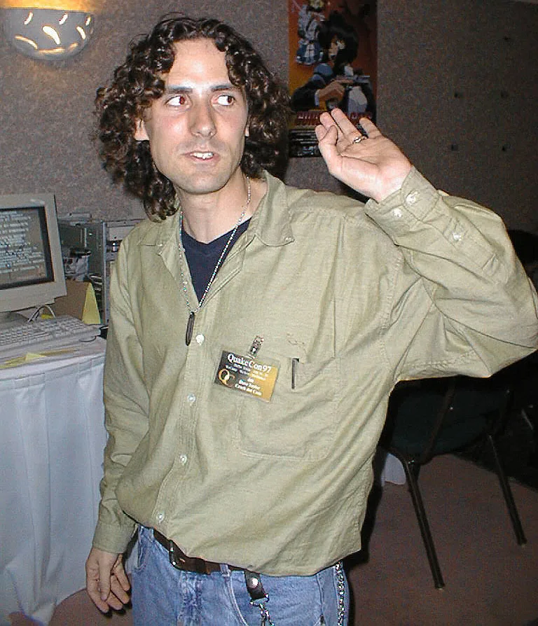 Dave Taylor in a green shirt at QuakeCon 1997, with a Crack dot Com badge on his chest and a raised hand