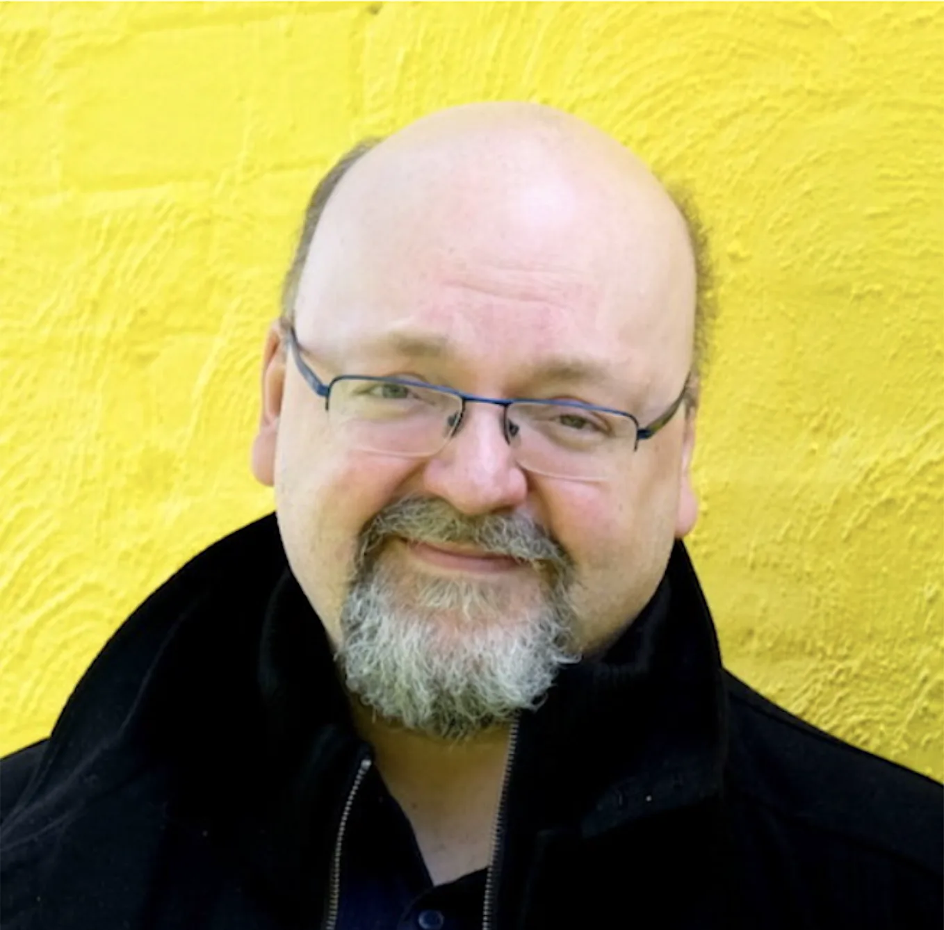 Portrait of David Gaider, a bearded man in glasses and a black top, against a yellow background