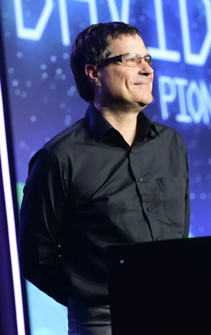 David Braben in a dark shirt on stage at the Game Developers Choice Awards, smiling, in front of a lit blue screen