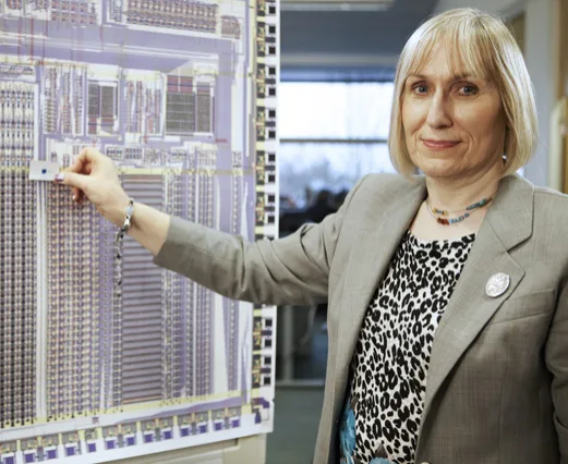 Sophie Wilson standing beside a large printed plan of a processor chip, pointing at it