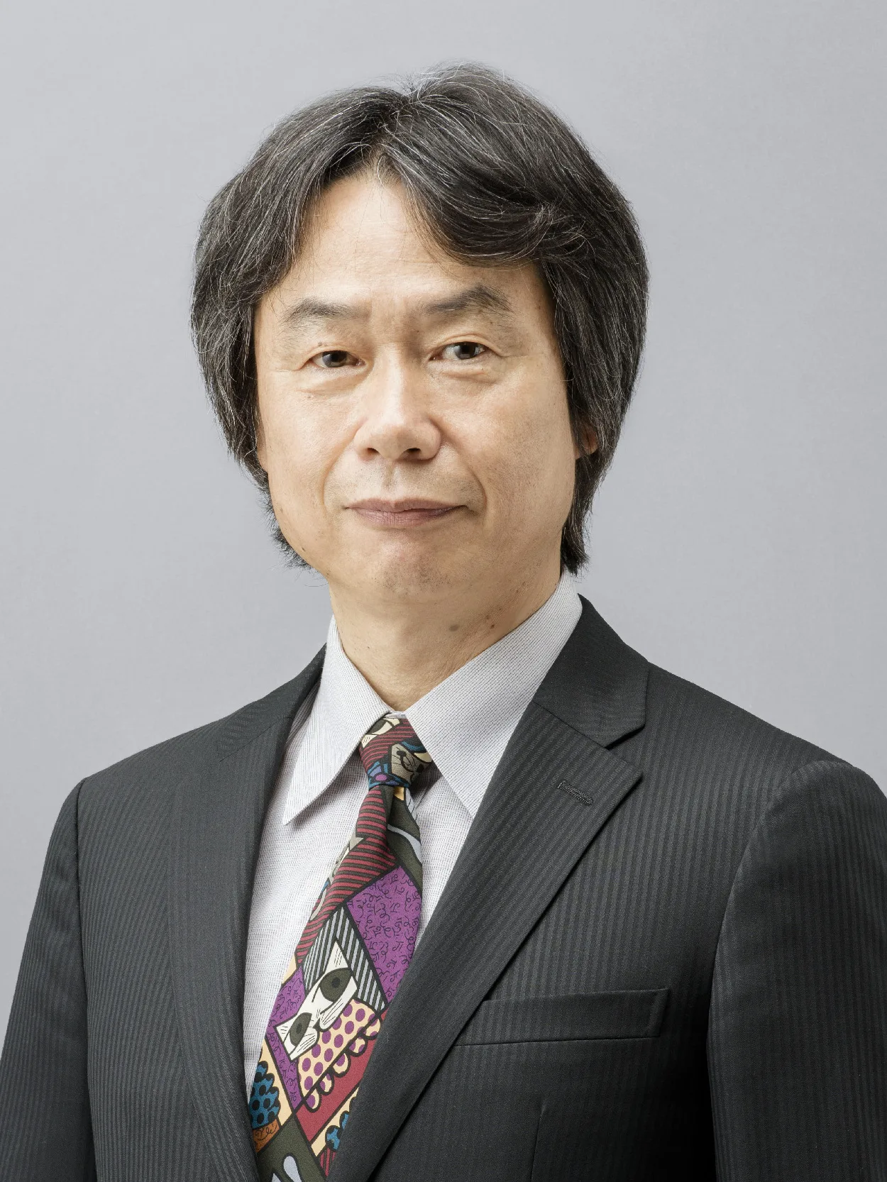 Portrait of Shigeru Miyamoto in a dark suit and a colorful patterned tie against a gray background