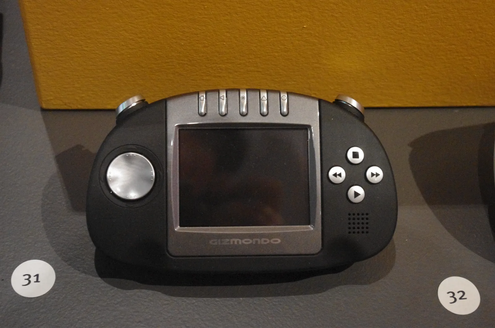 Black Gizmondo handheld with a small square screen, a silver stick on the left and four media buttons on the right