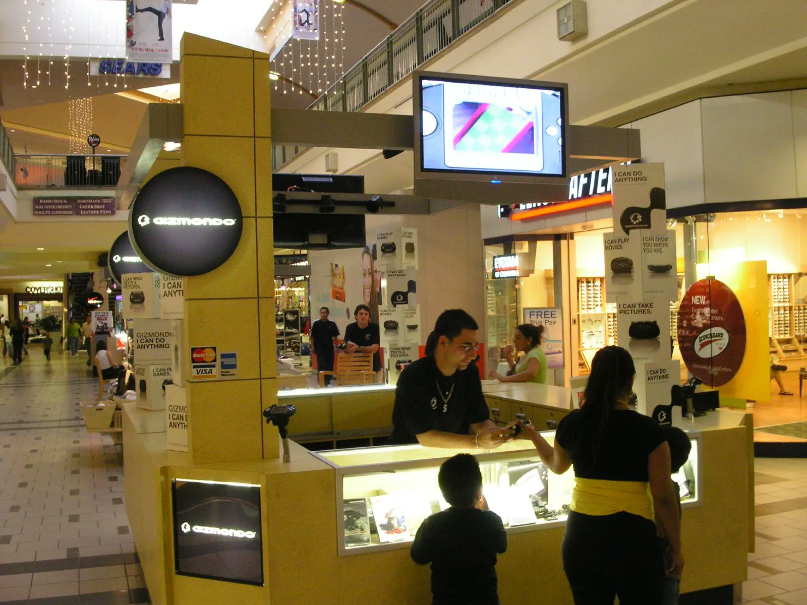 Yellow Gizmondo kiosk in a shopping mall with a staff member handing a console to a customer