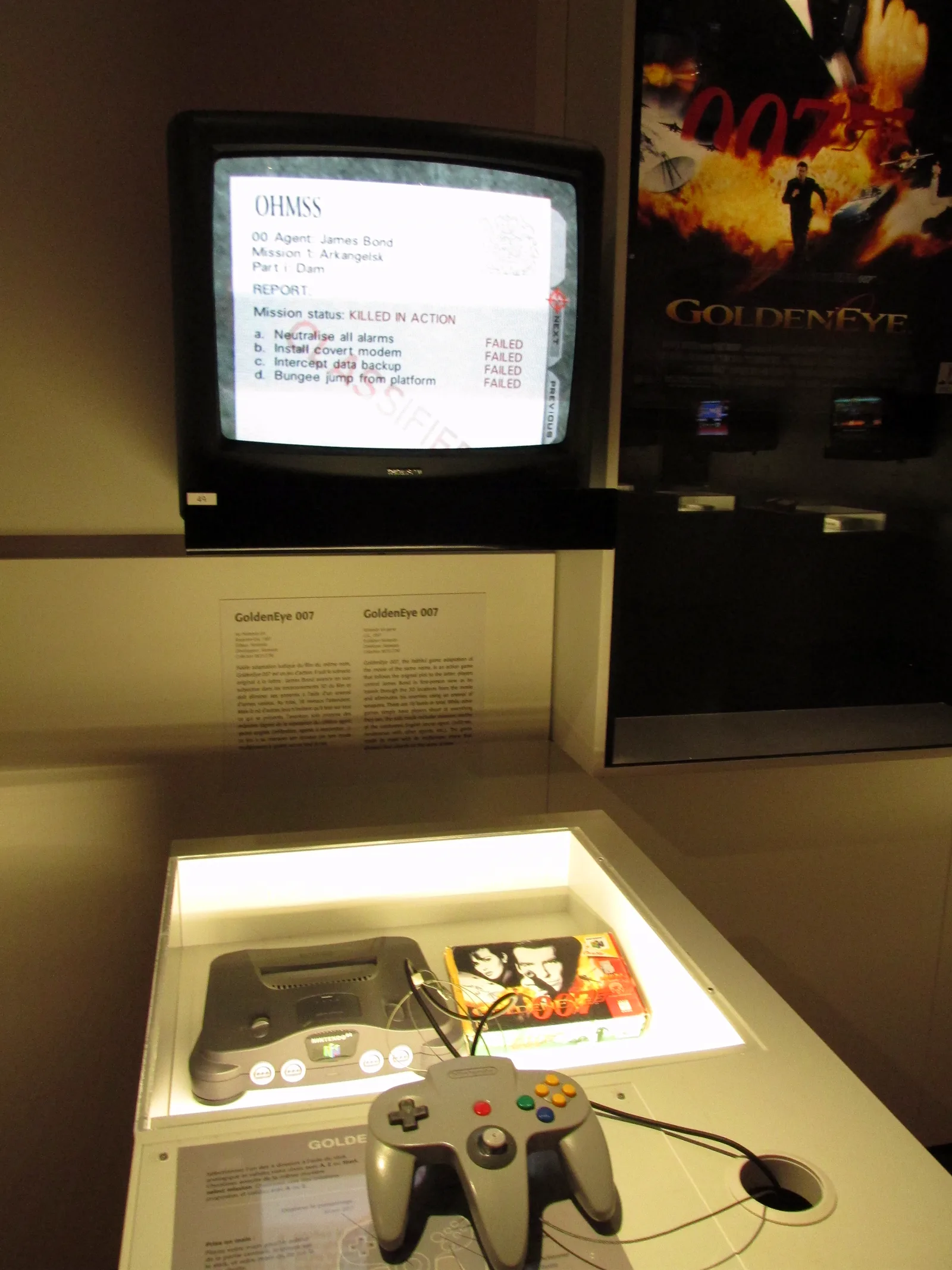 A Nintendo 64, a controller and a GoldenEye 007 box in a museum case under a TV showing a mission report