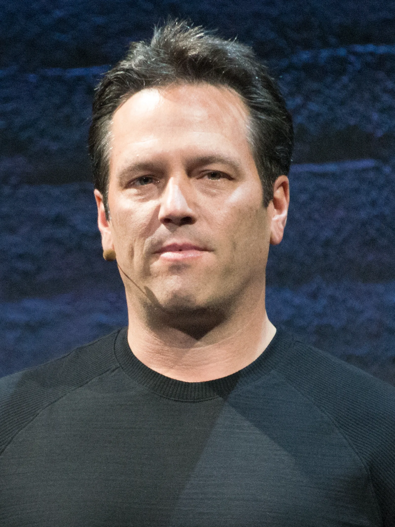 Close-up portrait of Phil Spencer on a stage, wearing a headset microphone and a dark sweater