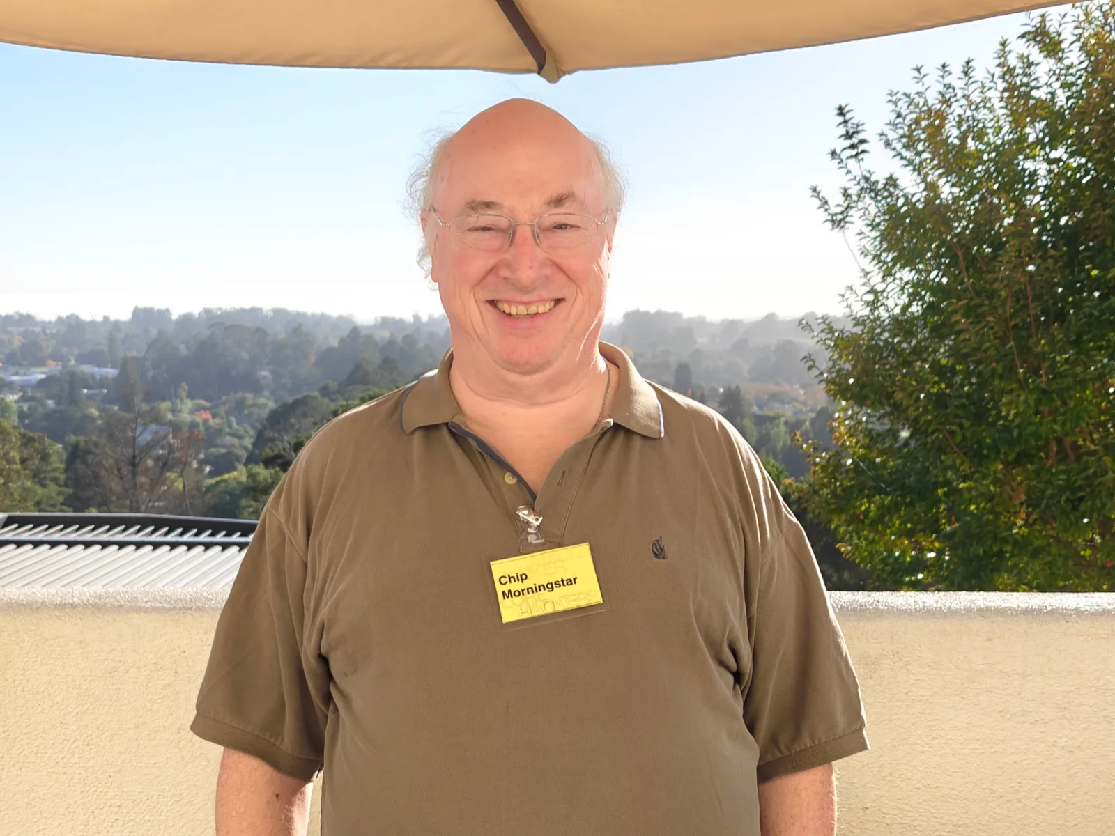 Chip Morningstar smiling on a terrace under an umbrella, wearing a name badge, with hills behind him