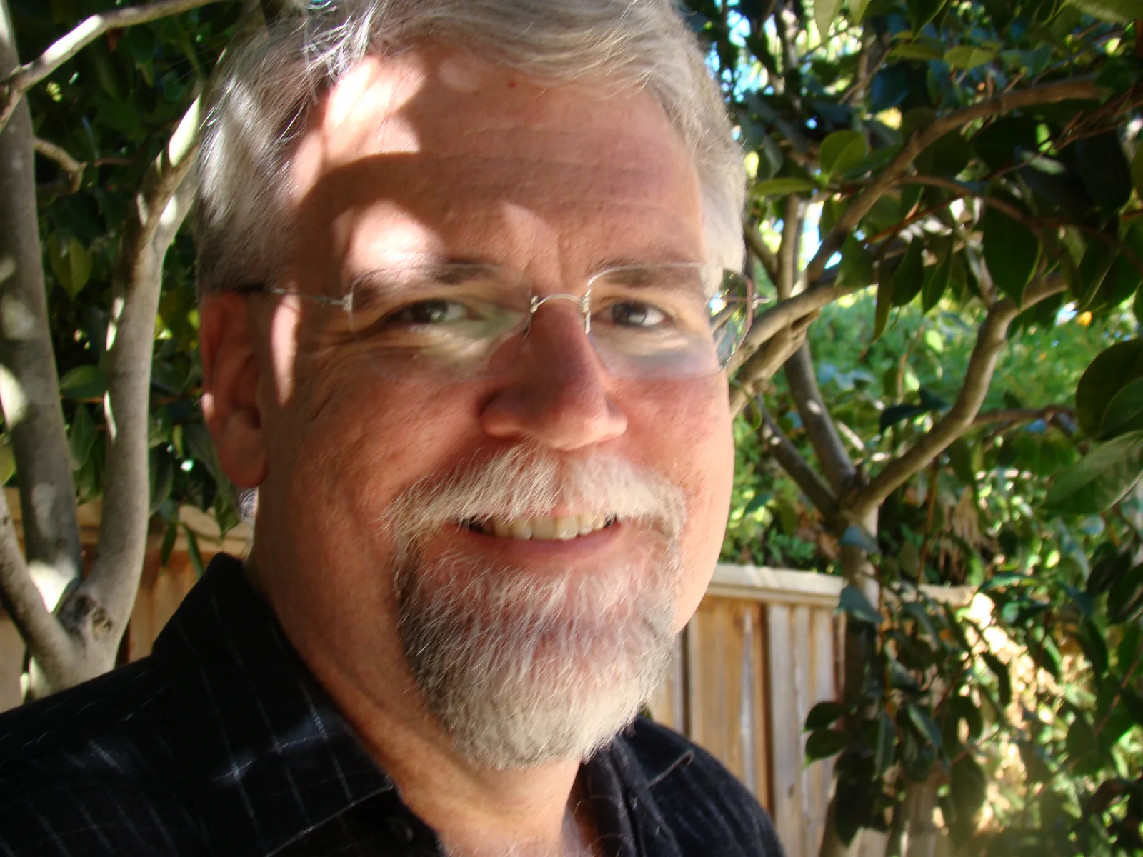 Close portrait of Randy Farmer, smiling, with gray hair, glasses and a goatee, outdoors in dappled sunlight