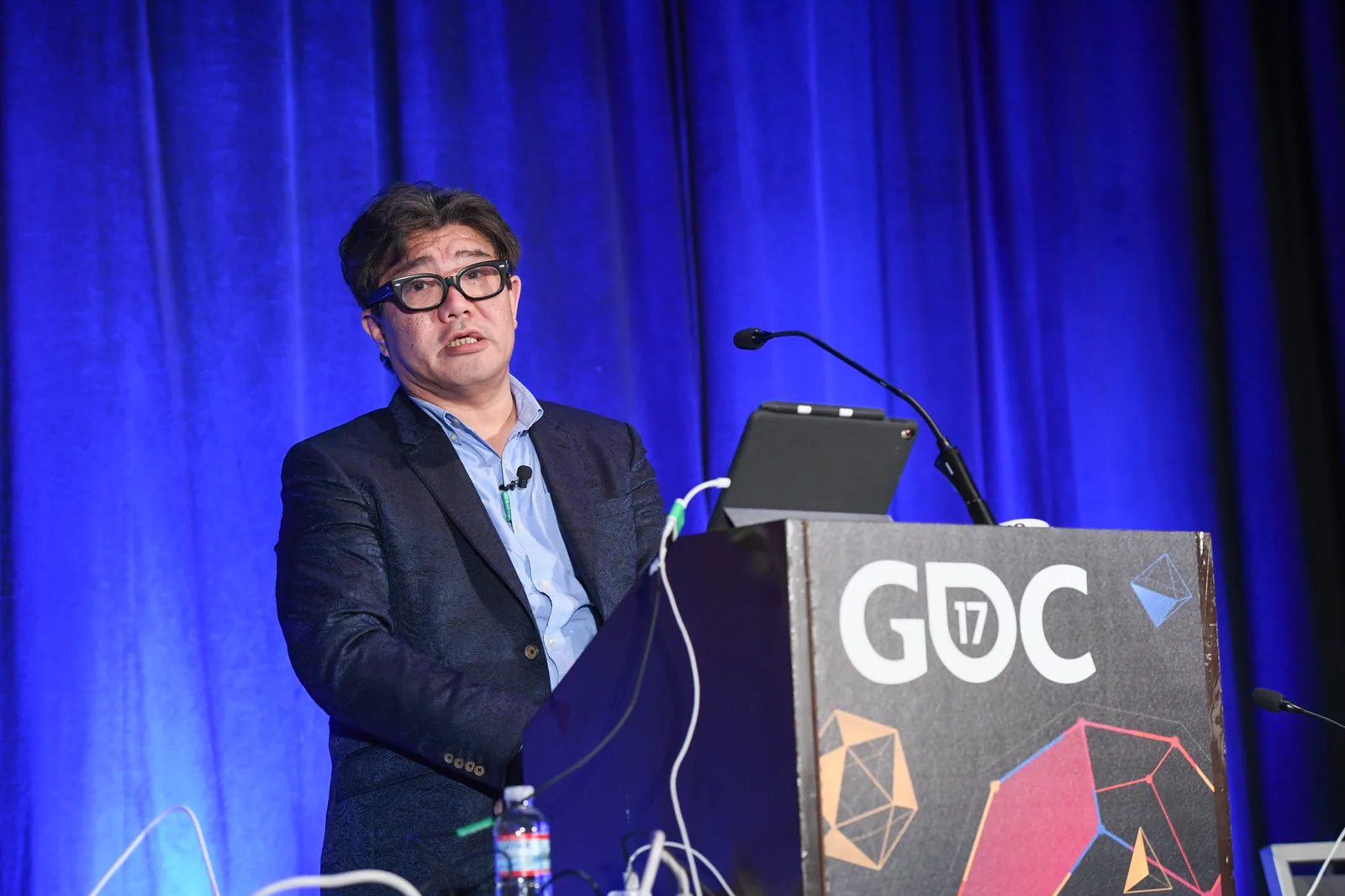 Yoot Saito speaking at a lectern marked GDC 17 in front of a blue curtain